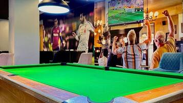 A pool table with overhead lighting, a television displaying a sports event, and a bar area with patrons.