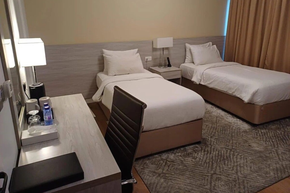 A hotel room with two beds, a desk with a lamp, a chair, and a small table with a bottle and tissues.