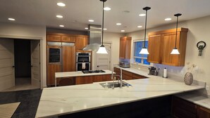 Een moderne keuken met houten kastjes, een marmeren eiland, roestvrijstalen apparatuur, hangende lampen en uitzicht op het groen door een raam.