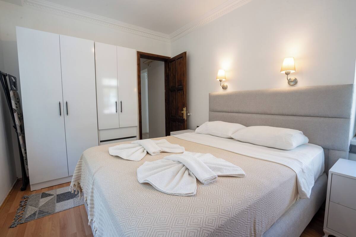 A neatly made bed with towels, a wardrobe, a bedside table, and two wall-mounted lamps.