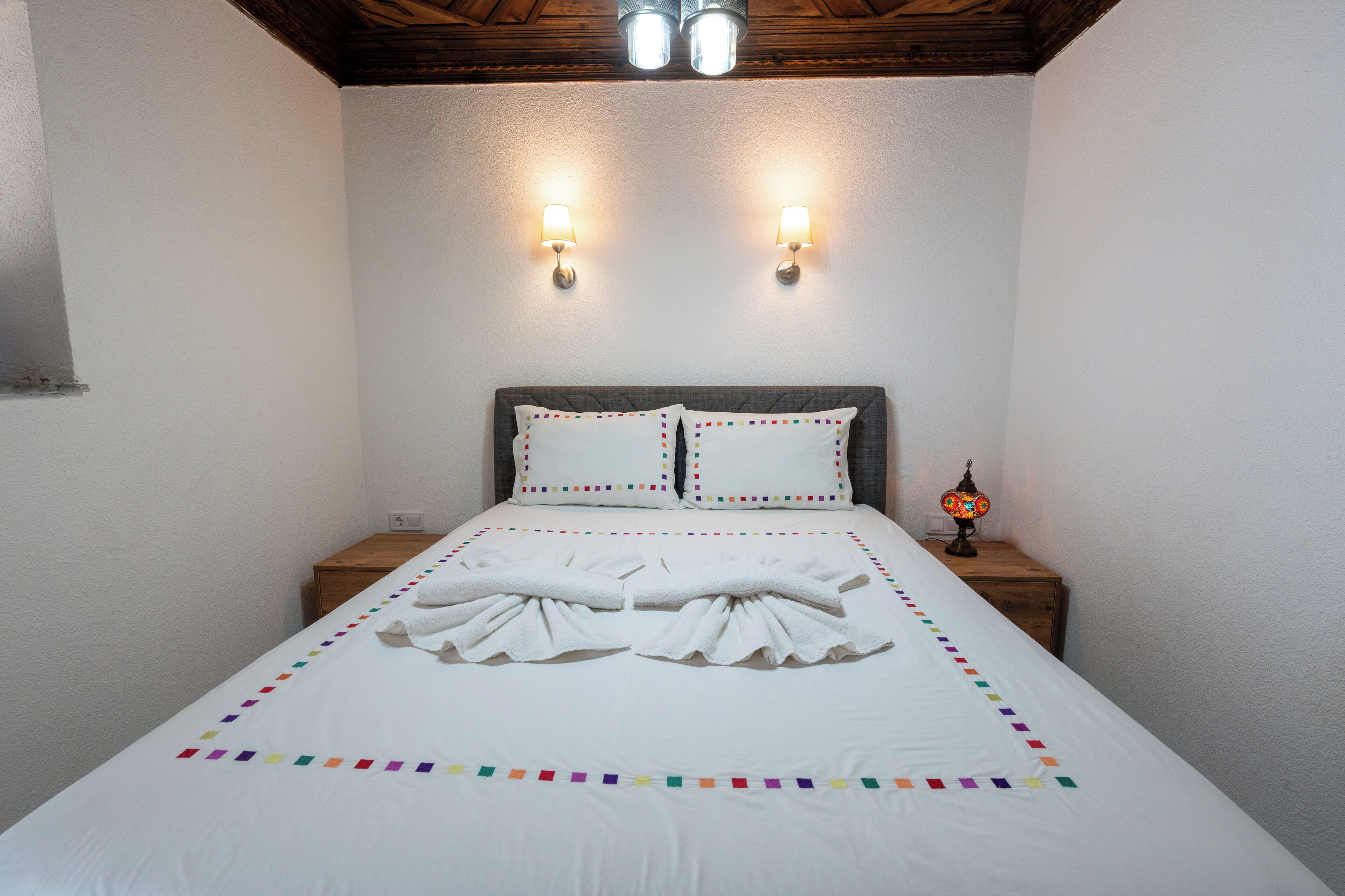 A neatly made bed with white linens and colorful pillows, two wall-mounted lamps, and a small table with a lamp beside the bed.