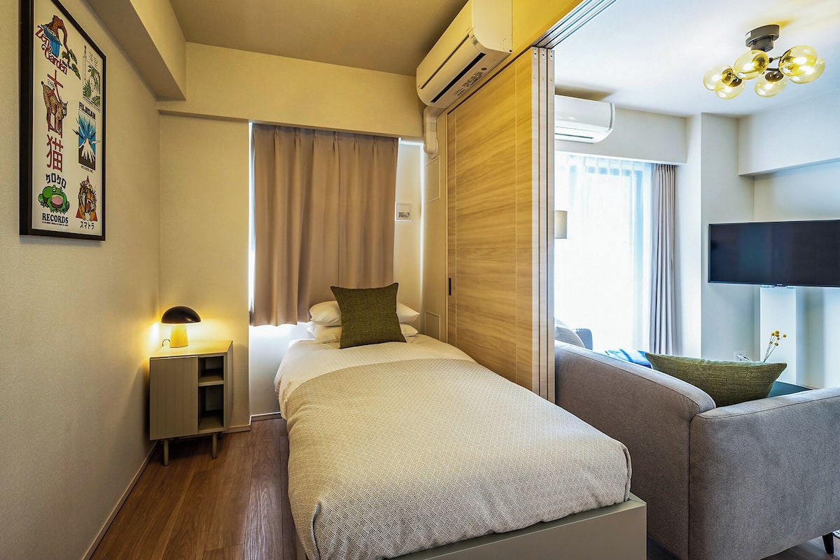 A hotel room with a bed, a bedside table, a lamp, an air conditioning unit, a TV, and a window with curtains.