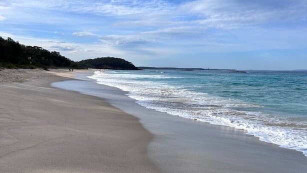 Una extensa platja de sorra amb onades suaus, cel blau i una terra a la distància.