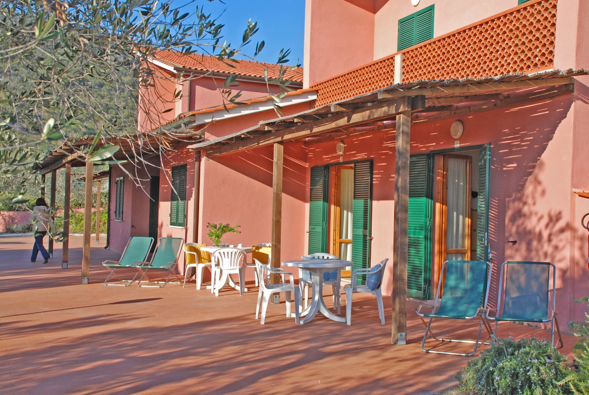 Ein Gebäude mit roter Fassade, grünen Fensterläden und einer überdachten Terrasse mit Gartenmöbeln.