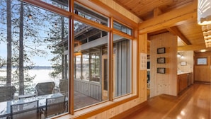 Interior de madera con grandes ventanales que dan a un lago, una zona de comedor con mesas al aire libre y una cocina.