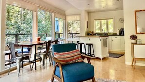 A spacious living area with a dining table, chairs, and a blue armchair. A kitchen with a bar counter and large windows is visible in the background. - Redwood Treehouse with Hot Tub (Guerneville)