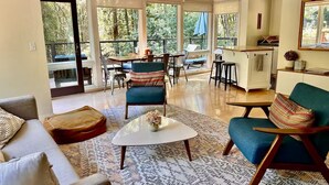 A spacious living area with a sofa, armchairs, a coffee table, and a dining area with chairs and a table. Large windows provide views of the outdoors. - Redwood Treehouse with Hot Tub (Guerneville)