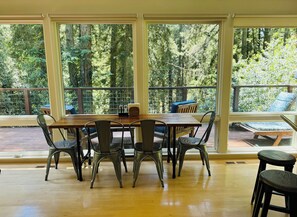 Un comedor con mesa de madera y sillas metálicas, ventanas amplias con vista a un bosque y un balcón con mobiliario exterior.