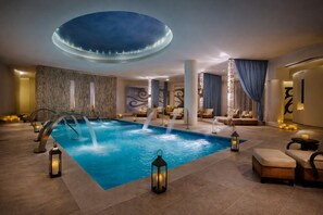 Ein großzügiger Innenpoolbereich mit einem zentralen Becken, mehreren Liegestühlen und stimmungsvoller Beleuchtung.