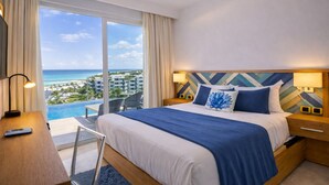 A hotel room with a bed, a desk, a chair, a nightstand, a lamp, and a view of the ocean and buildings. - Penthouse at Hard Rock Hotel (Punta Cana)