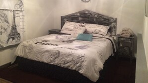 Une chambre à coucher avec un lit agrémenté de linge de lit sur le thème de Paris, une tête de lit ornée du logo de Paris et une table de chevet avec une lampe.