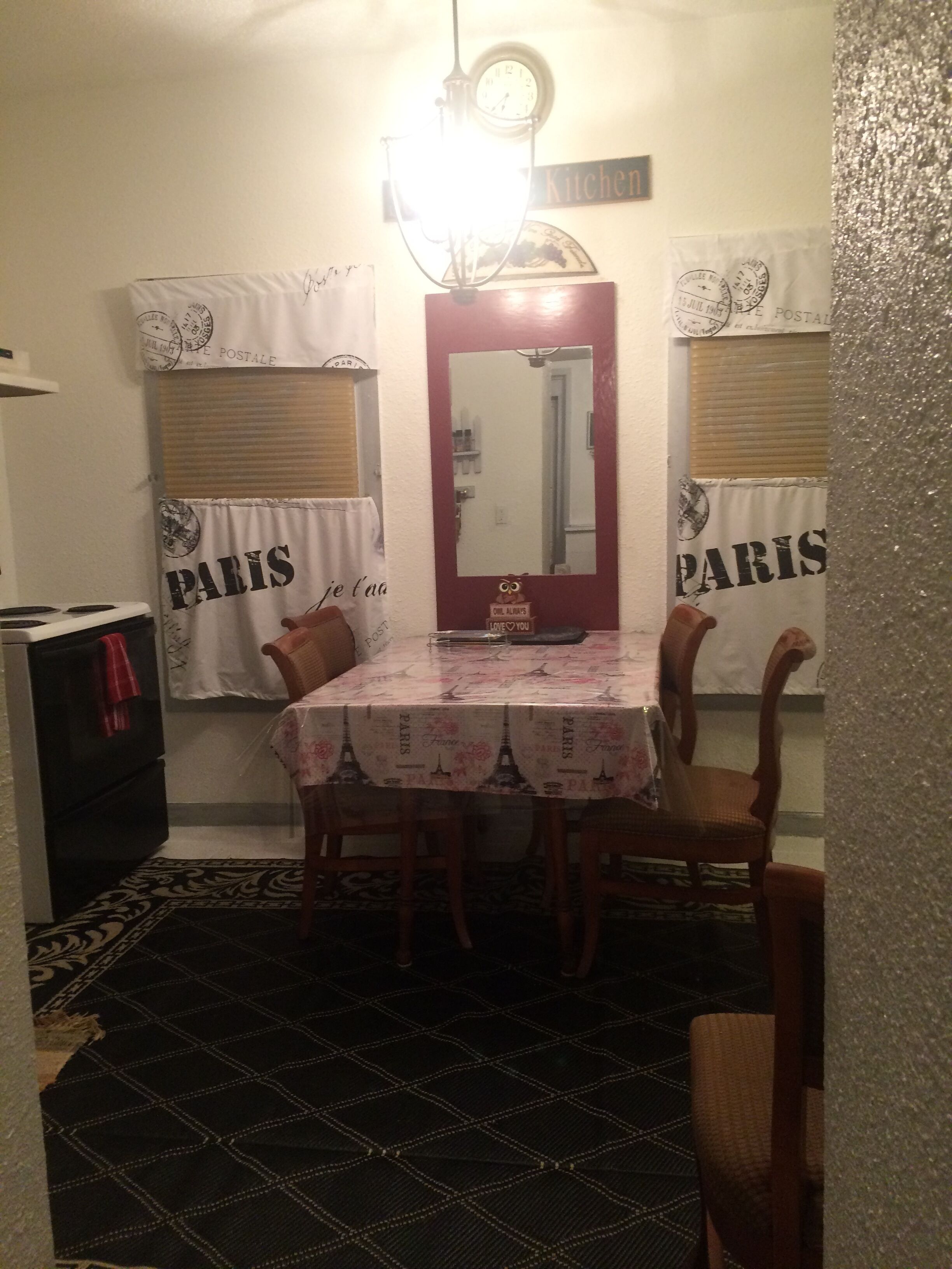 Uma área de refeições com uma mesa coberta por uma toalha com tema parisiense, ladeada por cadeiras. Um espelho está pendurado acima da mesa e o ambiente possui uma estética vintage, com um relógio na parede.