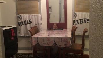 Uma área de refeições com uma mesa coberta por uma toalha com tema parisiense, ladeada por cadeiras. Um espelho está pendurado acima da mesa e o ambiente possui uma estética vintage, com um relógio na parede.