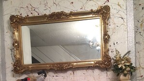 Une cheminée surmontée d’un miroir à cadre doré, un vase rempli de fleurs sur la droite et une étagère où sont disposés divers objets.