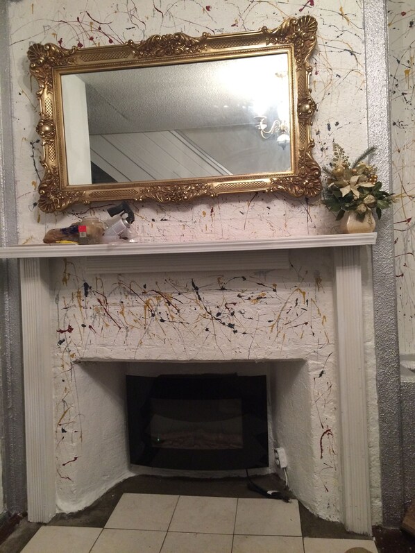 Una chimenea con un espejo dorado encima, un jarrón con flores a la derecha y un estante con varios objetos.