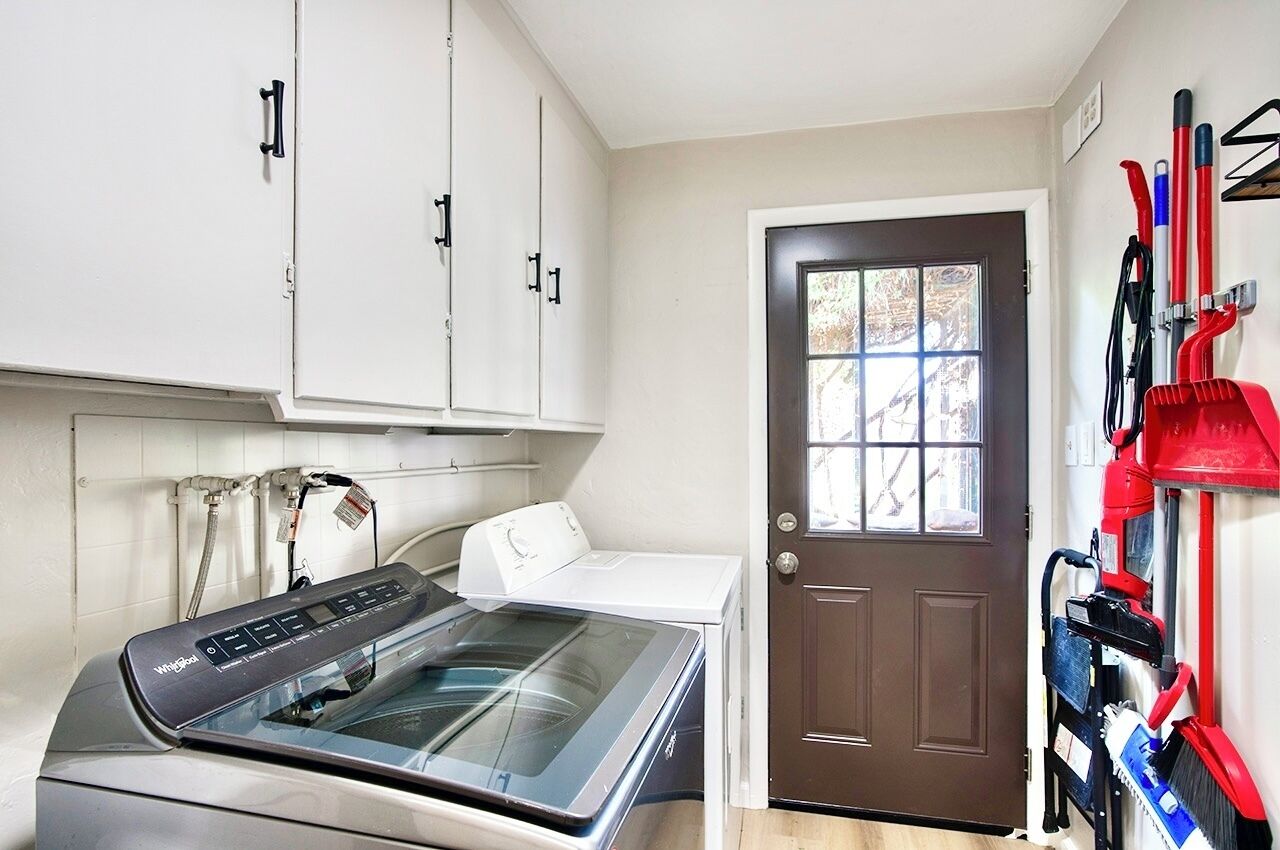 A laundry room with a front-loading washer and dryer, a brown door, and a wall-mounted tool rack with brooms and mops.