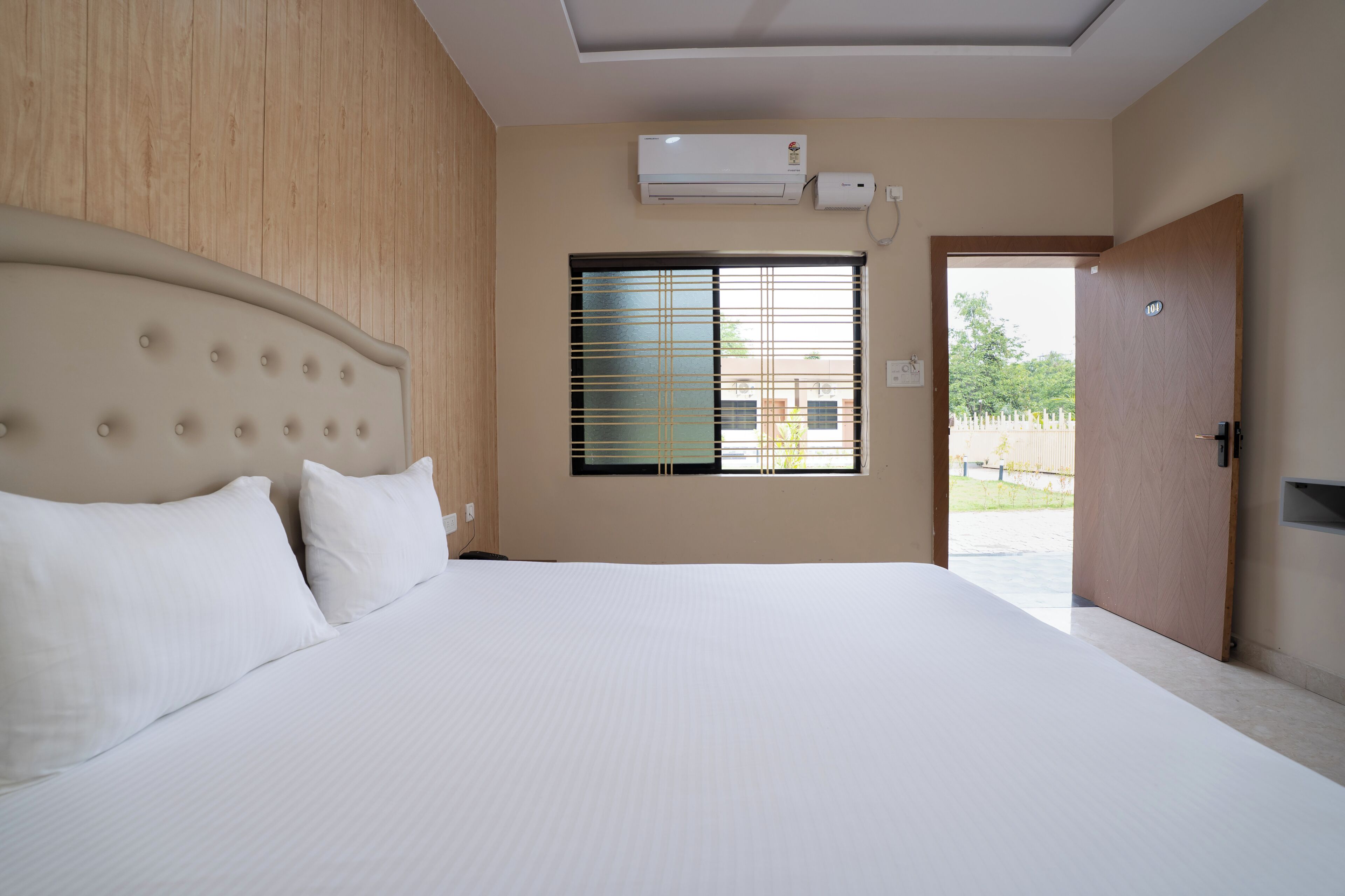 A hotel room with a large bed, an air conditioning unit, a window with blinds, and a door.