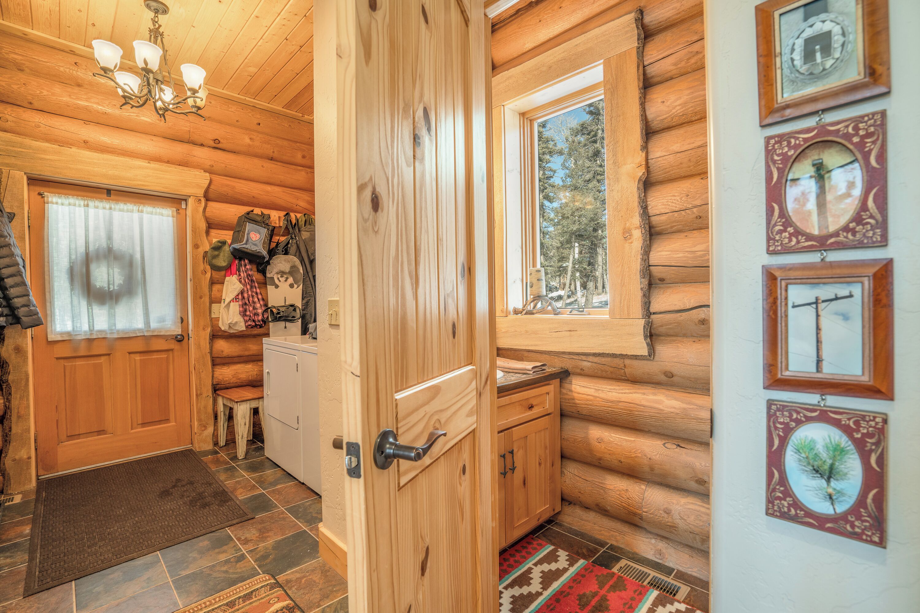 A wooden cabin interior with a wooden door, a bench, a small table, and a window with a view of trees.