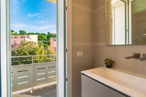 A balcony with a view of buildings and trees, a white bathroom with a sink and mirror, and a wall-mounted light switch. - Casa Sotto Bomba  a stone's throw from the sea. (Portoferraio)