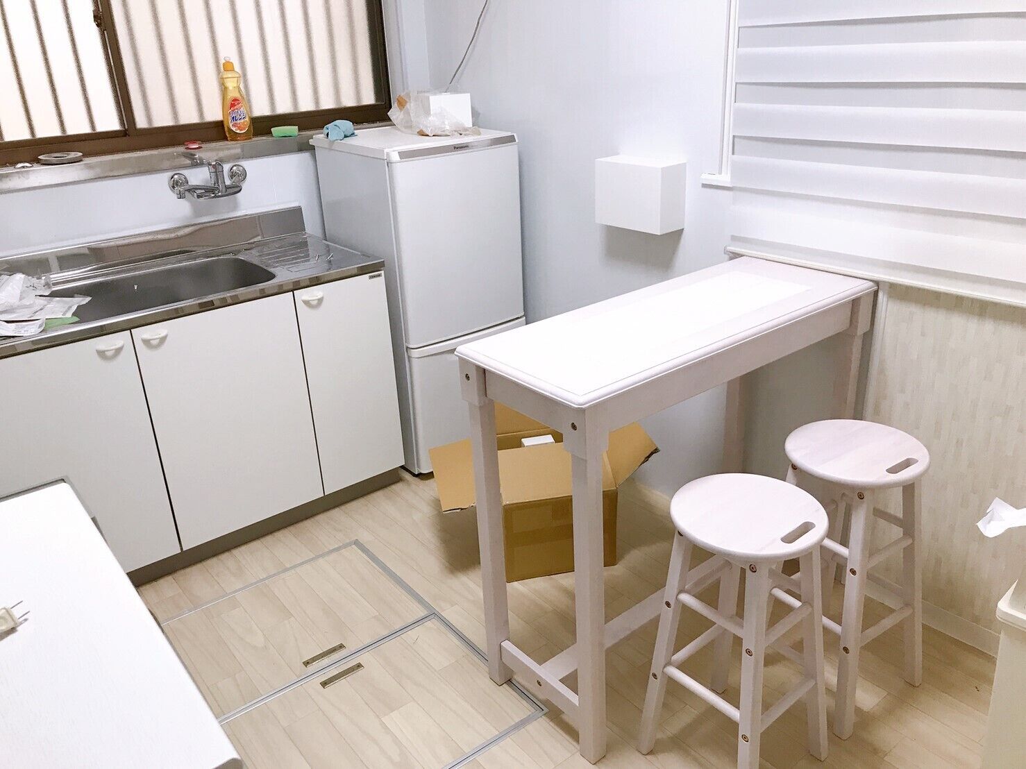 Una cocina compacta con un lavamanos, un refrigerador pequeño, una mesa blanca con taburetes y una ventana con persianas.