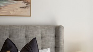 A neatly made bed with a grey upholstered headboard, two dark pillows, and a white pillow. A white bedside lamp is on a small round table beside the bed. - St Georges Gem—Spacious & Well-located Family Stay (Saint Georges)