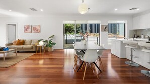 A modern living room with a sofa, coffee table, and plants. There is a dining area with a table and chairs. The kitchen has white cabinets and a sink. - St Georges Gem—Spacious & Well-located Family Stay (Saint Georges)
