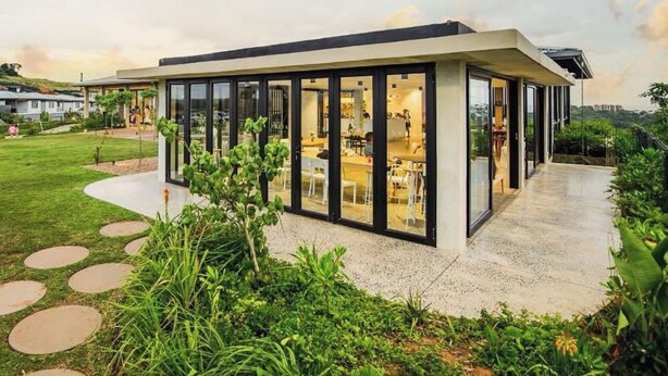 Un bâtiment moderne aux murs de verre, doté d’un patio et entouré de verdure, avec un ciel dégagé.