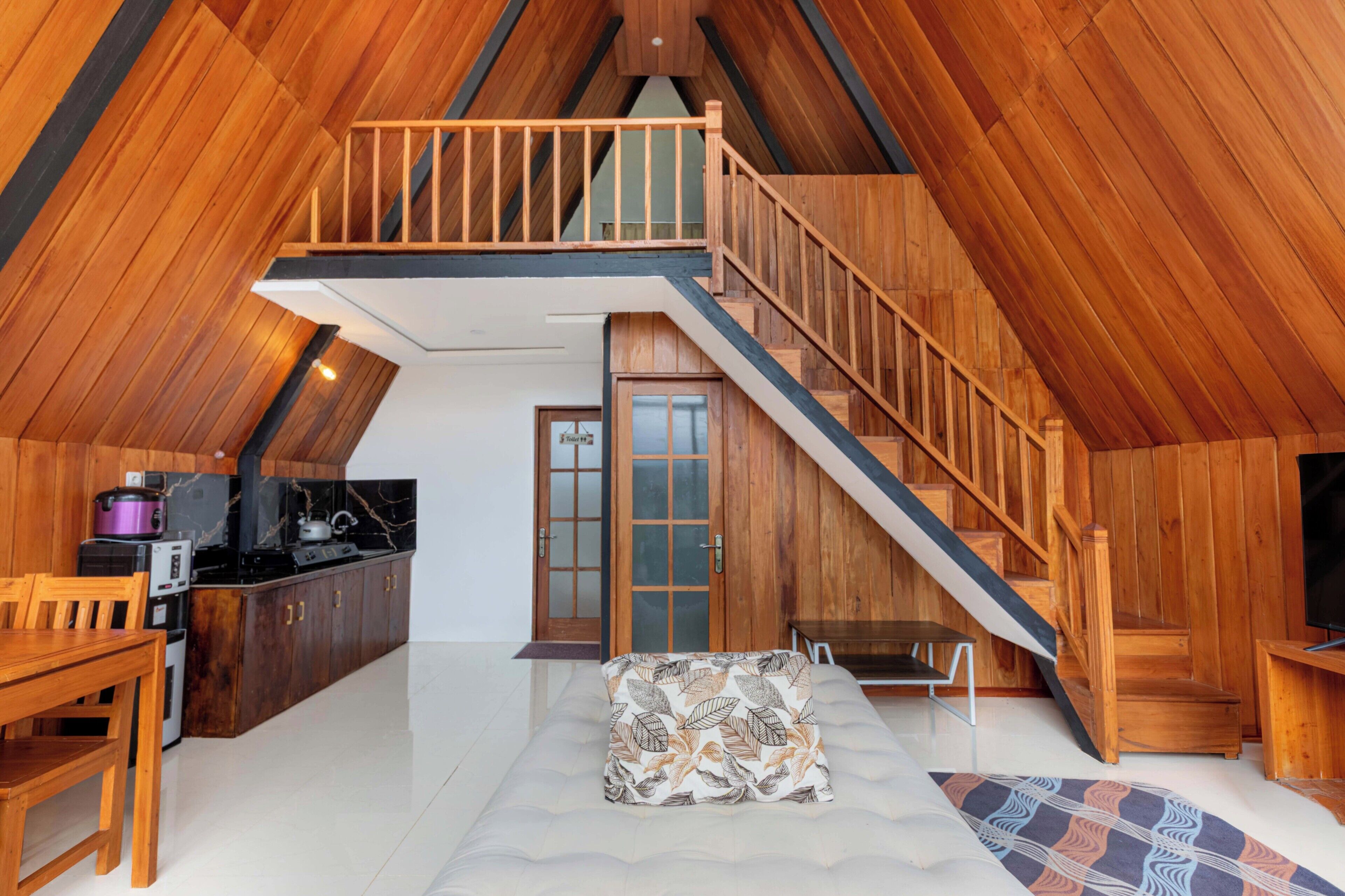 A loft-style room with a wooden staircase, a kitchen area, and a seating area with a patterned cushion.