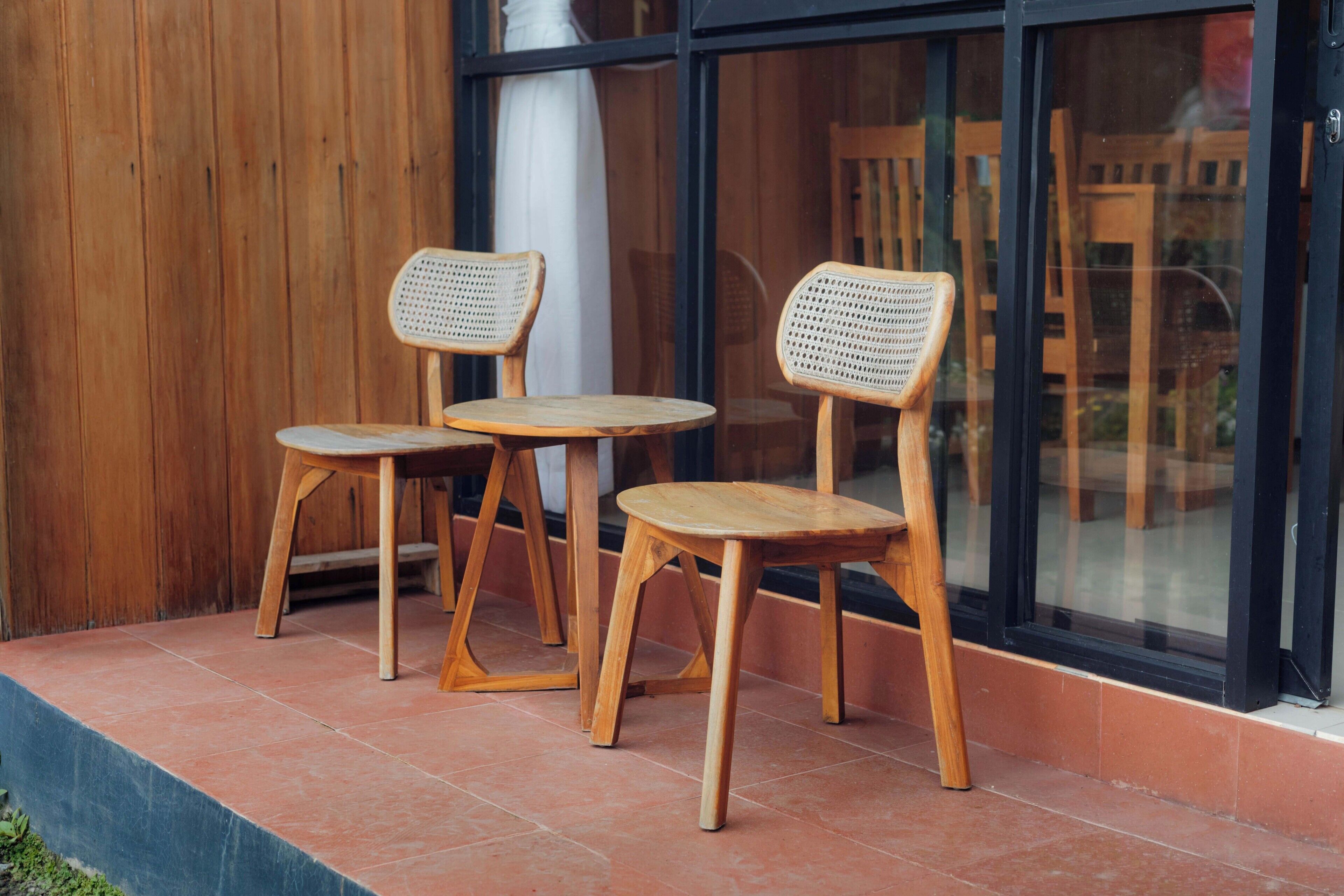 Two wooden chairs with woven seats and legs, placed on a tiled patio outside a building with large windows.