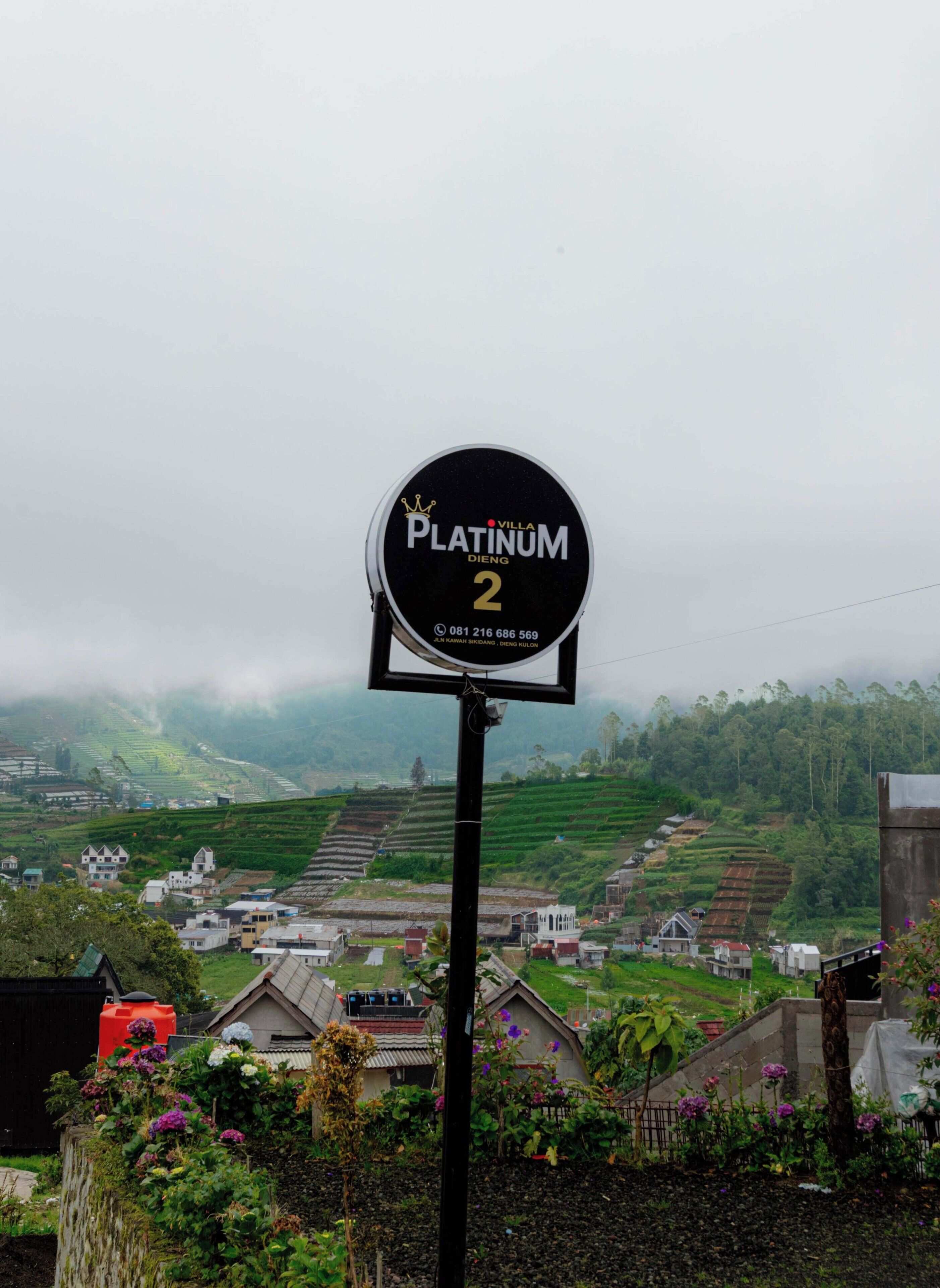 Schild für Villa Platinum DIENG 2 mit Blick auf gestufte Felder und Häuser im Hintergrund.