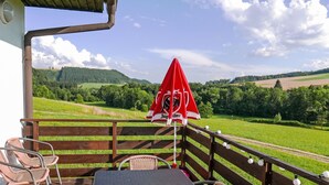 Een terras met rieten stoelen en een tafel, uitkijkend op een groen landschap met een rode parasol.