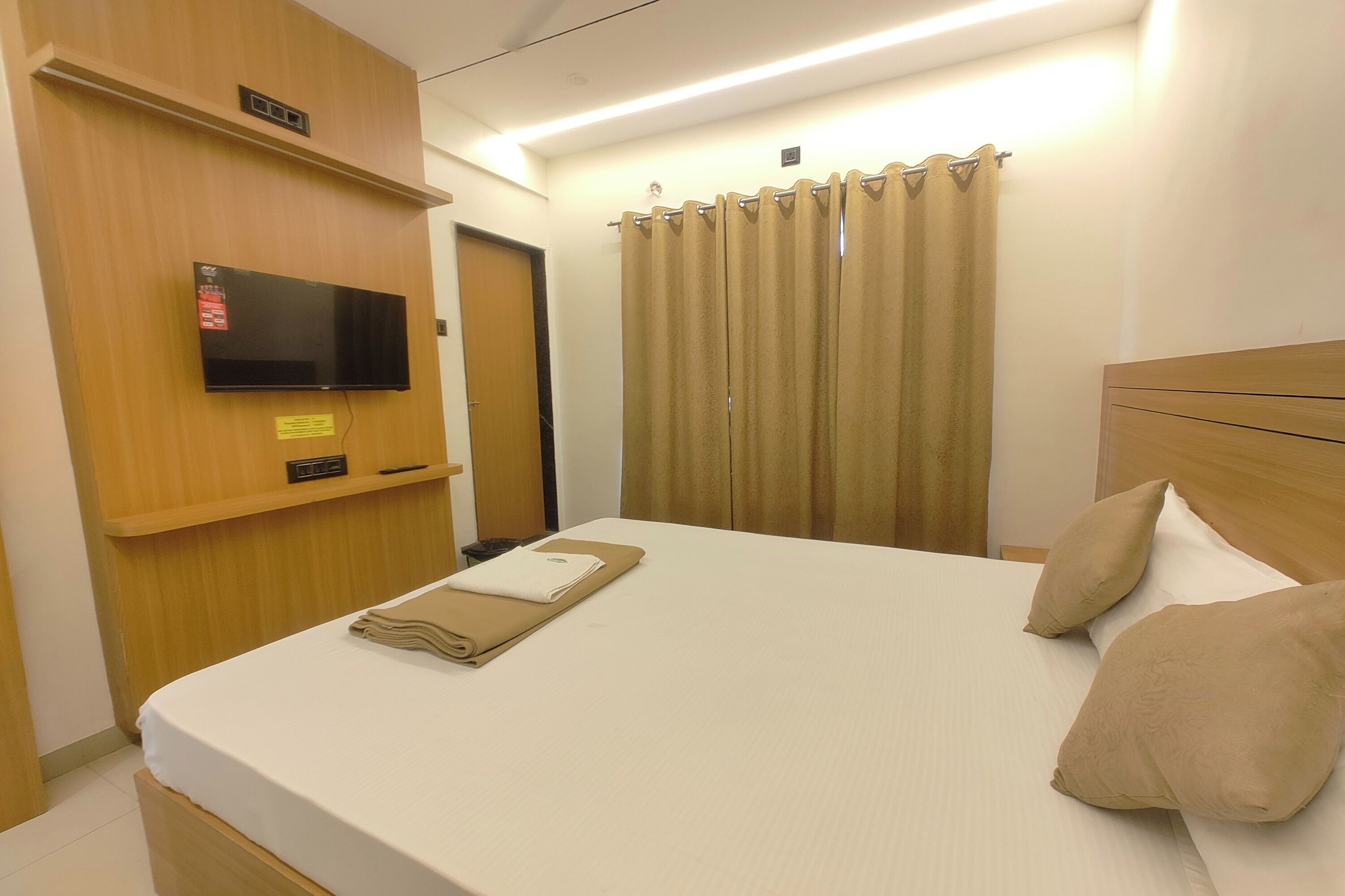 A hotel room with a bed, a television mounted on the wall, a curtain, and a wooden headboard.