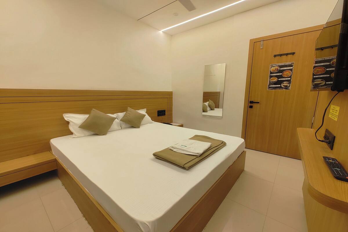 A hotel room with a wooden headboard, a neatly made bed, a bedside table with a remote control, and a door with a mirror and menu posters.