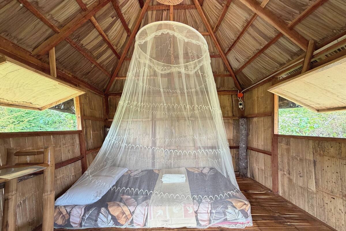 Una habitación de madera con una cama bajo una mosquitera, paredes de madera y techo de paja.