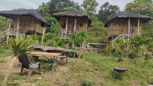 Tres casas del árbol de madera con techos de paja, rodeadas de vegetación y una pequeña mesa con una silla.