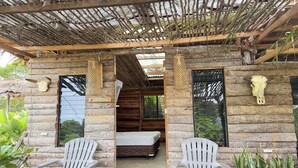 A rustic wooden cabin with a thatched roof, two white plastic chairs, a bed, and a small staircase leading to an upper level. - Tribu Kihud "Experience Simple Living" (San Juan)