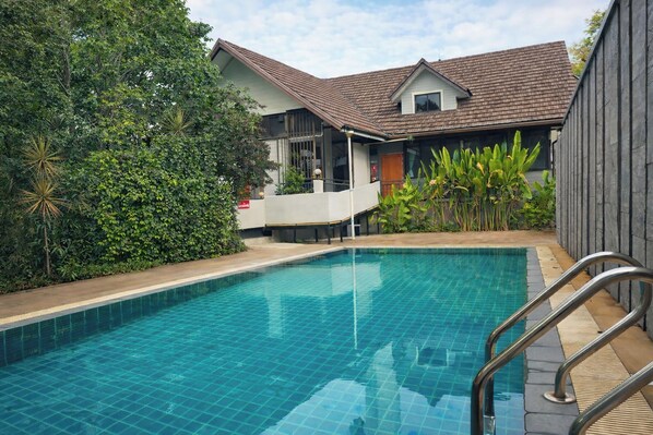 A swimming pool with a tiled deck, surrounded by lush greenery and a house with a tiled roof.