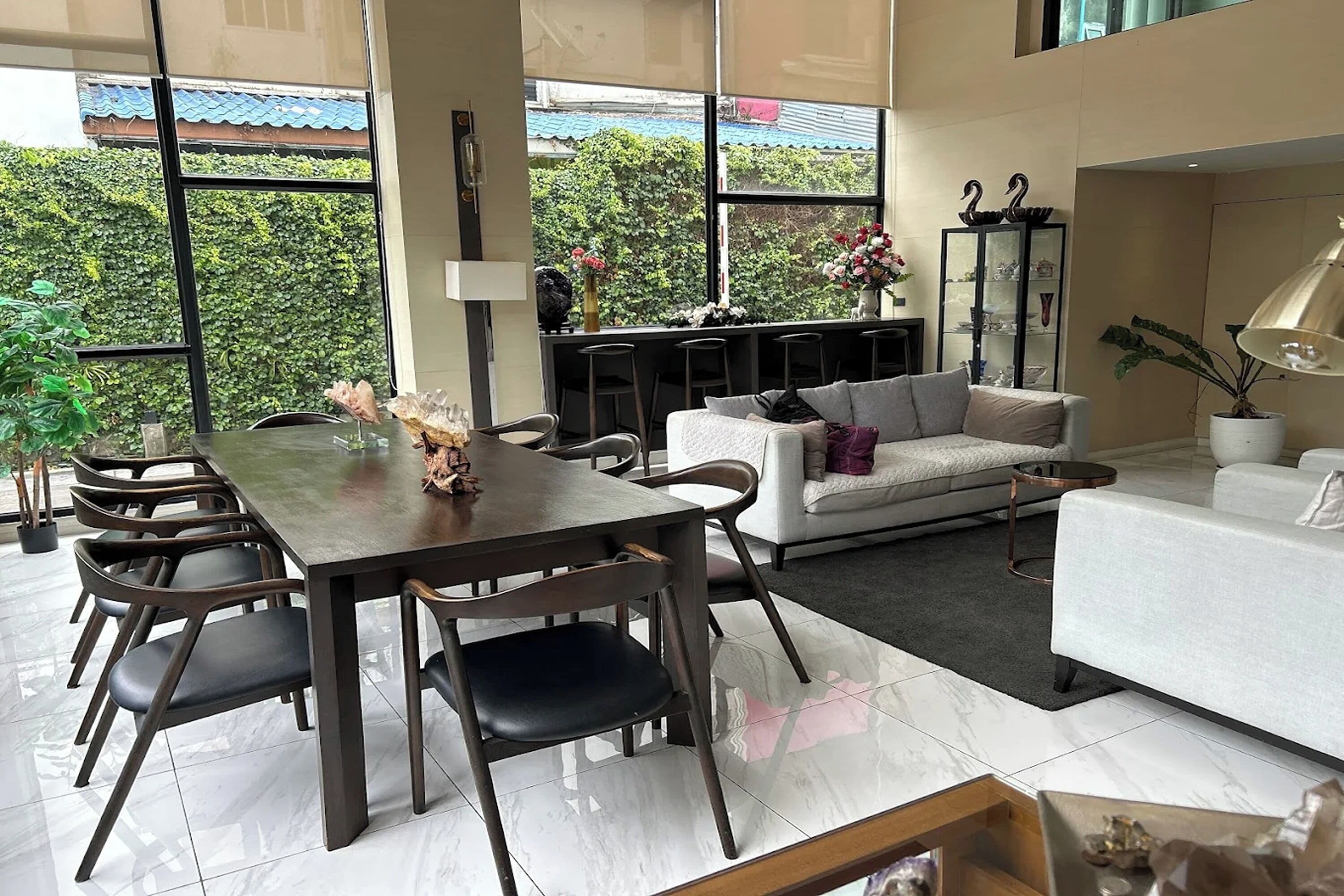 A modern living room with a dining area, large windows, and a view of greenery outside.