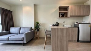 Una sala de estar moderna con un sofá gris, una pequeña mesa de comedor con sillas, una zona de cocina con refrigerador y un armario de madera.