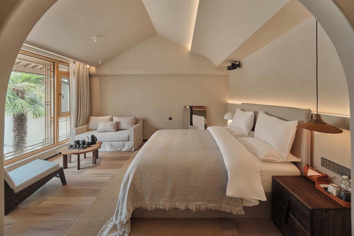 A spacious bedroom with a large bed, a sitting area with a sofa and table, and a view of greenery outside.