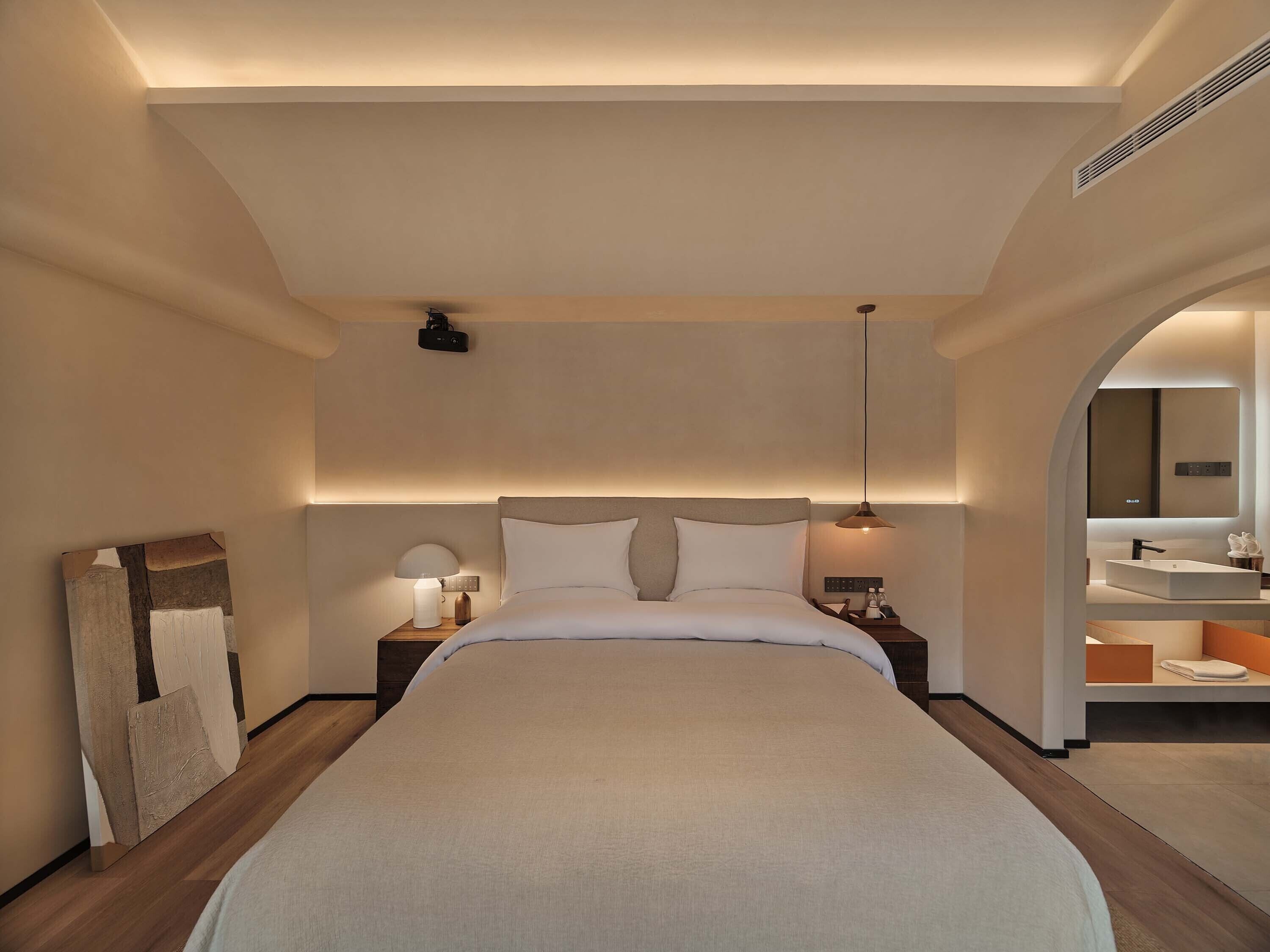 A modern hotel room with a large bed, bedside lamps, a ceiling projector, and a bathroom with a large mirror and sink.