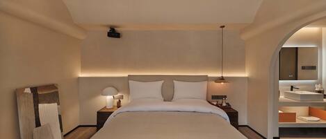 A modern hotel room with a large bed, bedside lamps, a ceiling projector, and a bathroom with a large mirror and sink.