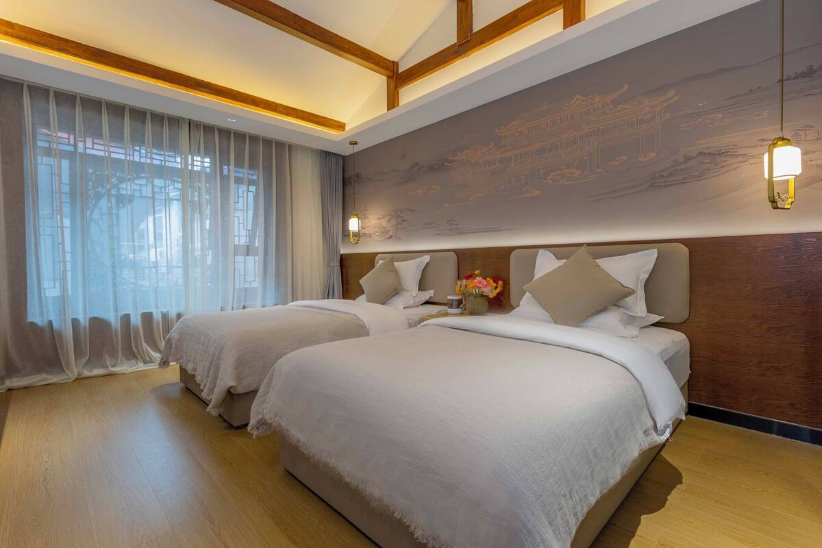 A hotel room with two beds, wooden flooring, a large window with sheer curtains, and a wall mural.