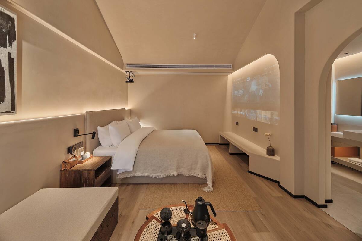 A modern hotel room with a large bed, a bedside table with a lamp, a small table with a kettle and cups, a projector screen on the wall, and a bathroom with a mirror and sink.