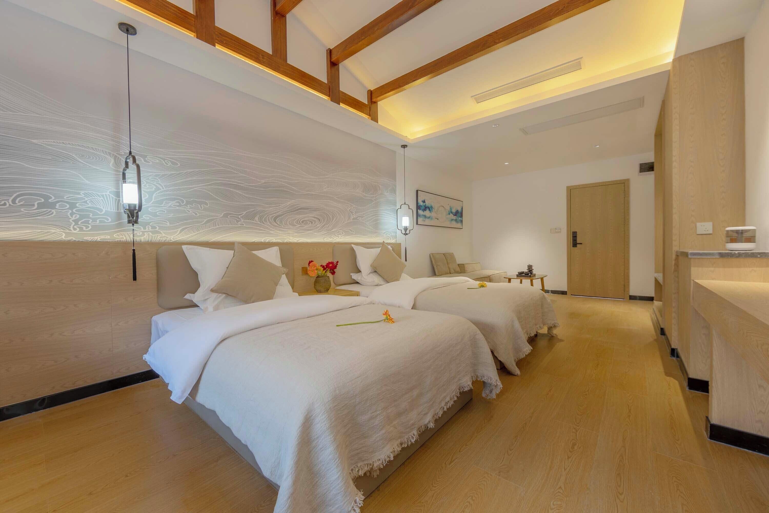 A hotel room with two beds, a wooden floor, a ceiling with exposed beams, a wall-mounted light fixture, and a small table with a vase of flowers.