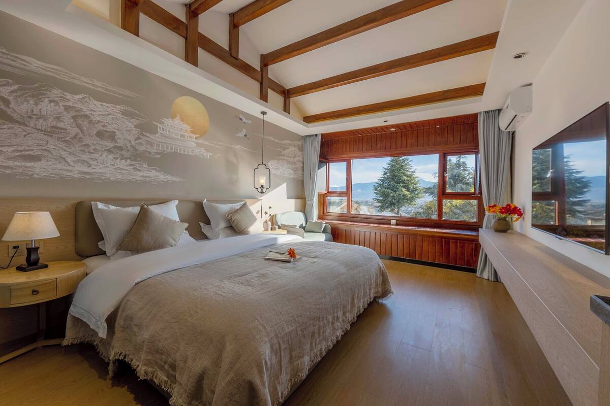 A spacious bedroom with a large bed, wooden beams on the ceiling, a nightstand with a lamp, and a window with a view of trees and mountains.
