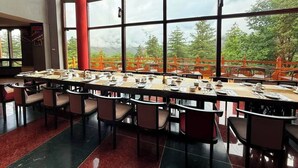 A dining area with a long table set for a meal, large windows with a view of greenery, and a red pillar. - Haven Resort and Spa (Paro)