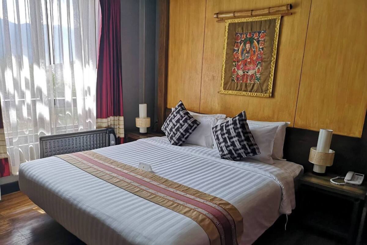 Habitación de hotel con una cama grande, dos mesitas de noche con lámparas, cabecero de madera, una ventana con cortinas y un cuadro en la pared.
