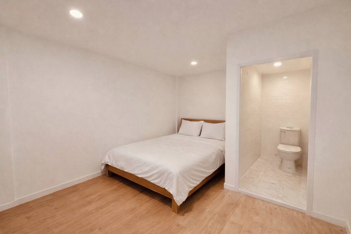 A bedroom with a wooden bed, white bedding, a white toilet, and a small bathroom.