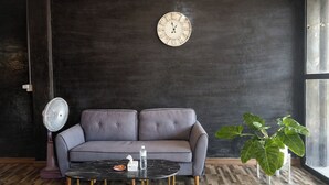 A modern room with a grey sofa, a round table with a water bottle, a clock, a fan, and a potted plant. - B&T Hostel (Chiang Rai)